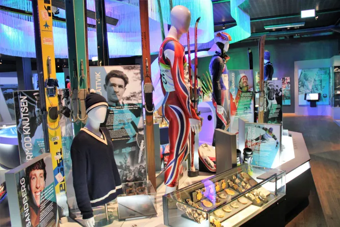 Innenraum einer Ausstellung mit Schaufensterpuppen in Skianzügen, stehenden Skiern, Infotafeln und Medaillen in Vitrinen..