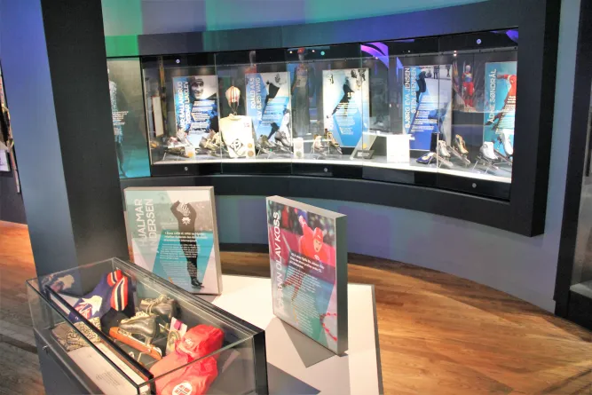 Innenraum einer Ausstellung mit gebogener Glasvitrine voller Schlittschuhe und Tafeln, davor weitere Exponate..