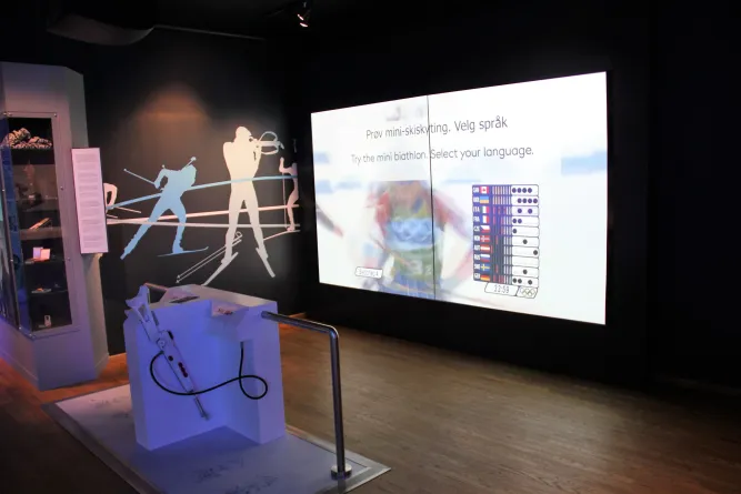 Dunkler Ausstellungsraum mit großem Bildschirm zur Sprachwahl für Mini-Biathlon und Gewehr-Attrappe am Podest..