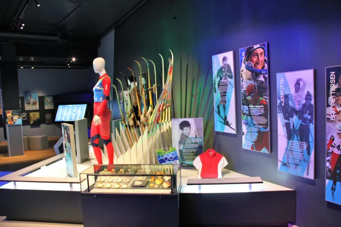 Ausstellungsraum mit Schaufenster voller Medaillen, Mannequin im Skianzug, vielen Skiern und Sportplakaten an der Wand..