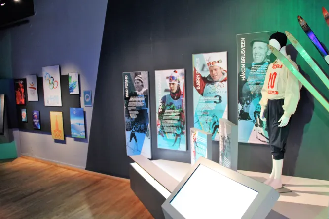 Innenansicht einer Ausstellung mit Sportpostern, Vitrinen und einer Schaufensterpuppe in Skikleidung samt Skiern..