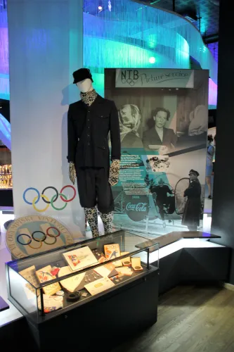 Mannequin in dunkler Kleidung neben einer Infotafel und Vitrine mit Souvenirs; im Hintergrund die Olympiaringe..