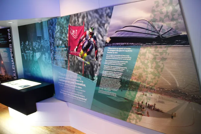 Schräg fotografierte Ausstellungswand mit Texttafeln, Skifahrer-Foto, Stadionaufnahme und beleuchtetem Display links..