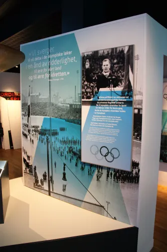 Große Museumstafel mit historischen Schwarzweißfotos, Textfeldern und den Olympischen Ringen in einem Innenraum..