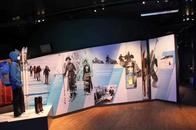 Innenansicht einer Museumsausstellung mit großen Wandtafeln zum Skisport und einer Skibekleidung auf Podest..