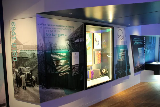 Ausstellungswand mit Texttafeln, Olympia-Symbolen und beleuchteter Vitrine in einem Museum; Holzboden im Vordergrund..