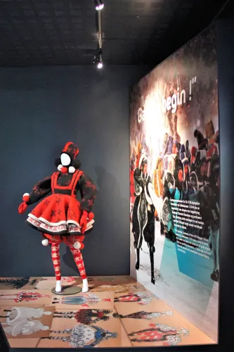 Rote und schwarze Puppenfigur in einem Museumsraum neben einer großen Wandgrafik mit Wintersportmotiven..