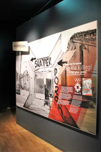 Große Museumstafel an dunkler Wand mit Foto einer Straße, Hinweisen „SNIPER“ und erläuterndem Textfeld..