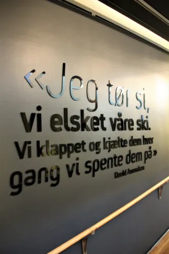 Schwarze und silberne 3D-Buchstaben mit norwegischem Zitat an dunkel gestrichener Innenwand, seitlich beleuchtet..
