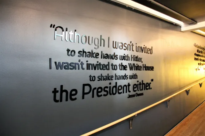 Schriftsatz-Zitat von Jesse Owens als Wandinstallation in einem Innenraumflur mit Geländer und Holzfußboden..