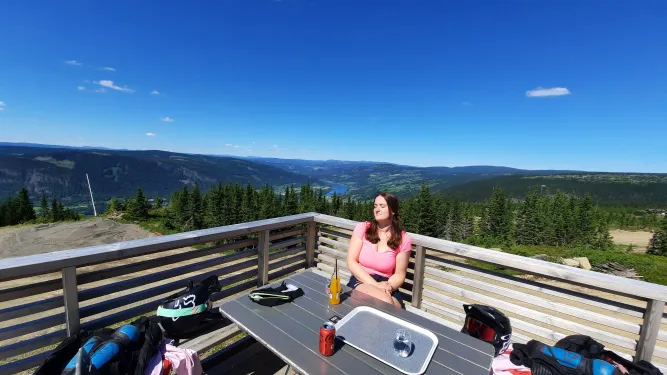 Eine Frau sitzt an einem Tisch auf einer Holzterrasse mit Getränken und Ausrüstung, dahinter Berge, Wald und blauer Himmel..