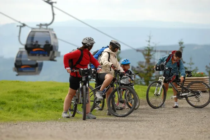 Vier mountainbikers met helmen staan op een grindpad bij een gondellift, met groene heuvels op de achtergrond..