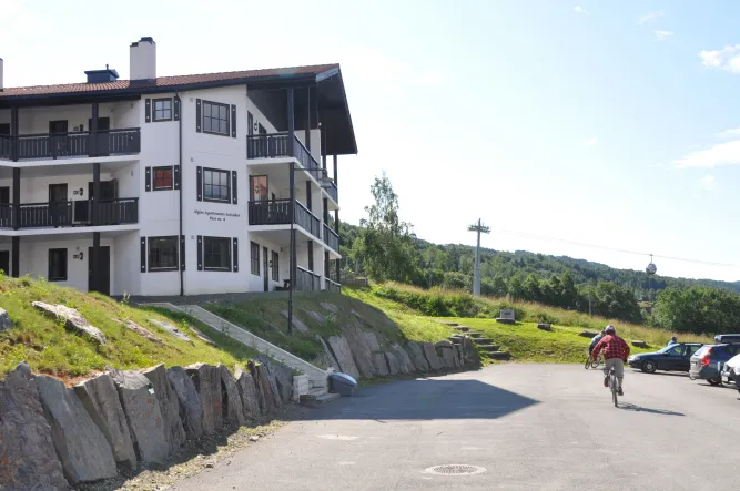 Een man fietst over een asfaltweg langs een wit appartementengebouw met balkons, grashelling en bomen..