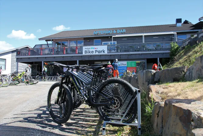 Rij mountainbikes in een rek voor een gebouw met 'Bike Park' en 'Afterski & Bar' onder een heldere blauwe lucht..