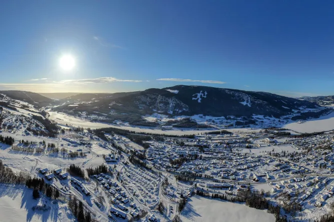 Luftaufnahme eines verschneiten Tals mit Wohnsiedlung, Fluss und bewaldeten Bergen unter tief stehender Sonne..