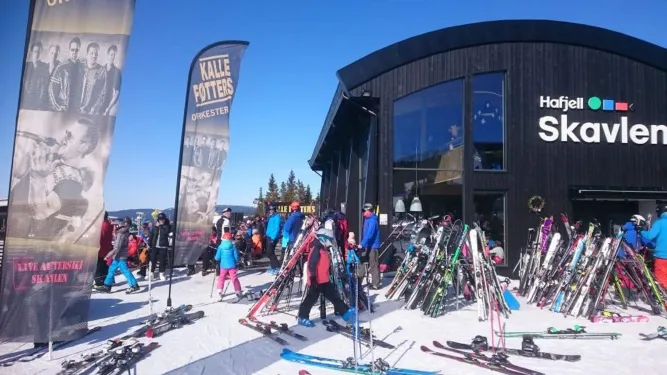 Viele Skifahrer stehen vor einem dunklen Gebäude, daneben Reihen abgestellter Ski und zwei Banner im Schnee..