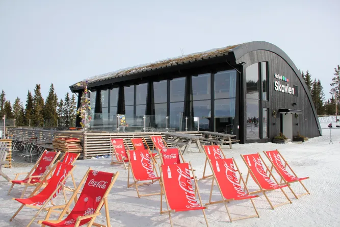 Moderne Berghütte mit großen Fenstern und geschwungenem Dach; davor rote Liegestühle auf verschneiter Terrasse..