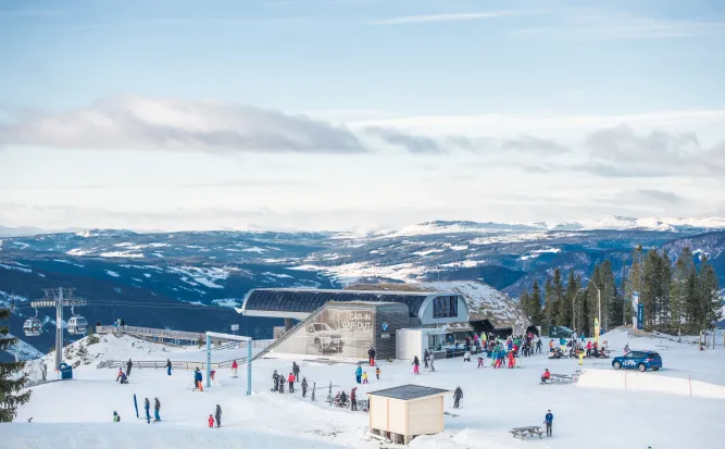 Verschneite Skipiste mit vielen Skifahrern an einer Liftstation, Bergen im Hintergrund und blauem Himmel..