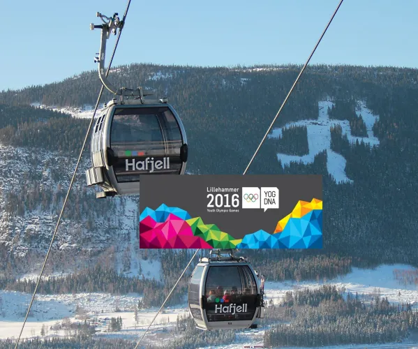 Zwei Gondelkabinen der Seilbahn schweben vor einem verschneiten Bergwald bei blauem Himmel; mittig ein Banner..
